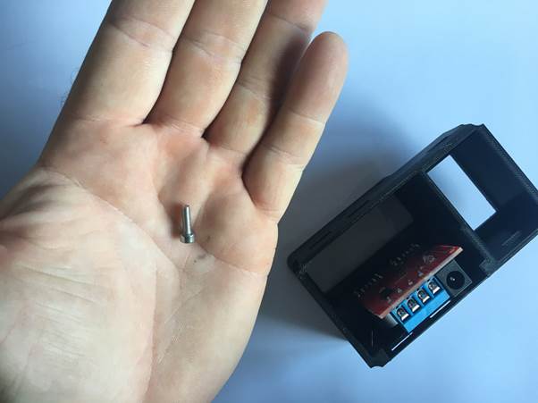 A hand holding a small retaining screw next to a black 3D printed clock housing with an electronic module inside.