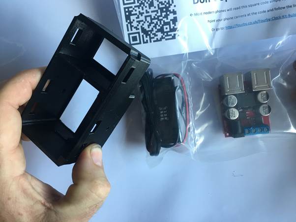 A person holding a black plastic housing for a USB clock kit, with a USB module and power cables visible in a sealed bag and a QR code notice in the background.