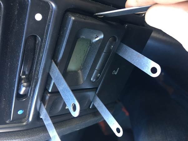 A hand using a tool to remove a clock from a car dashboard, with shims positioned around the clock to assist in its removal.