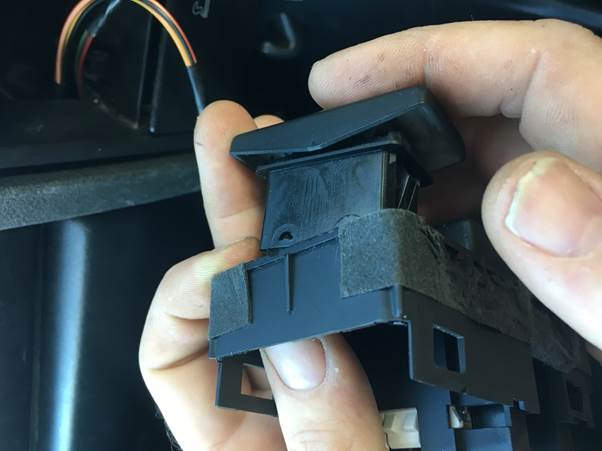 A person holding a black switch component while installing it into a panel in a car, with electronic wires visible in the background.